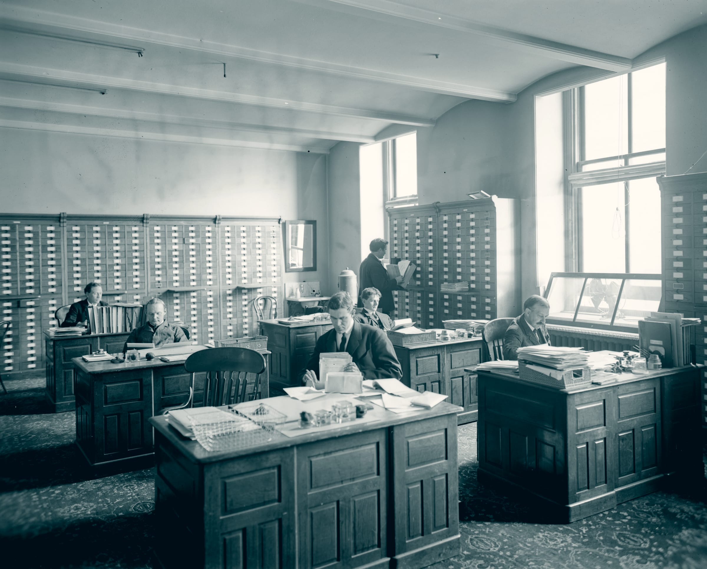 The records room of the United States Patent Office in 1907: clerks at wooden desks stacked with paper and wire in-trays, card-catalogue cabinets covering the walls floor to ceiling.